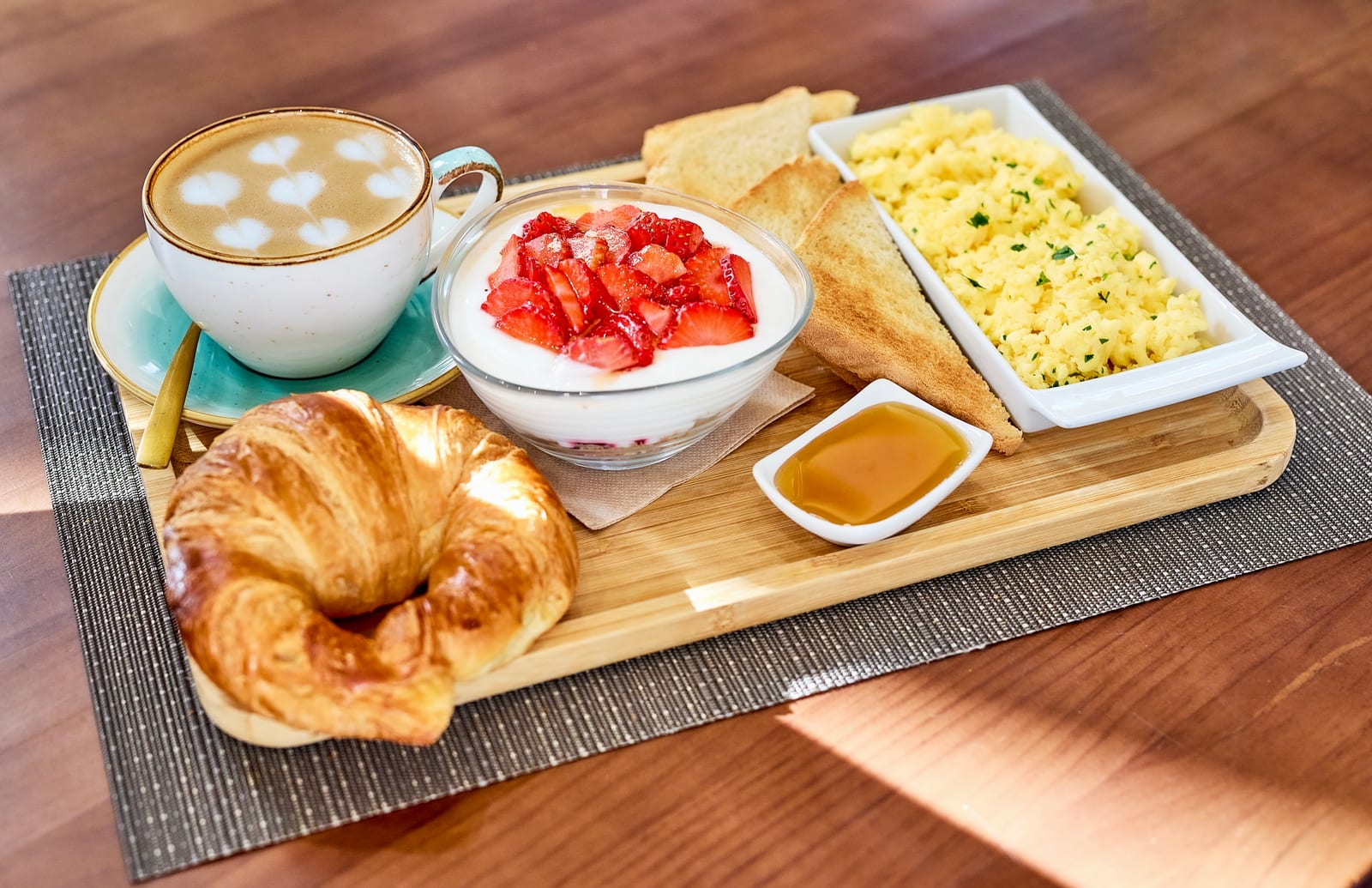 Breakfast board with a latte, croissant, yoghurt and strawberries, toast, honey and scrambled eggs